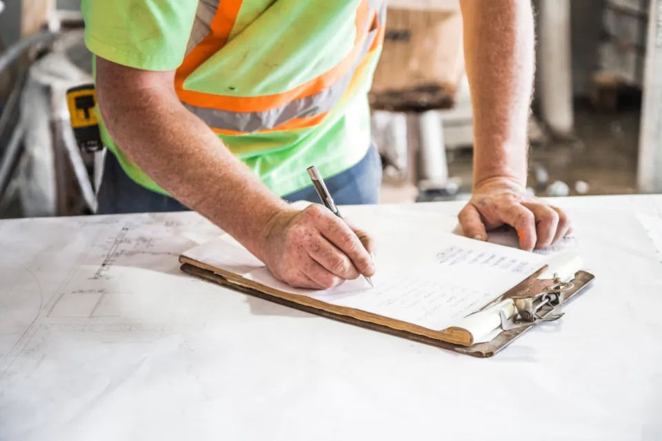 Worker in a high-visibility vest writing on a clipboard over drawings